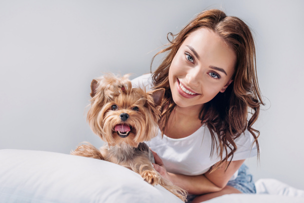 portrait of young smiling woman with yorkshire terrier resting on bed isolated on grey - Valokuva, kuva