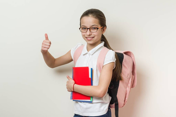 Girl school student, with a backpack and notebooks showing thumbs up sign, background bright wall in school - Фото, изображение