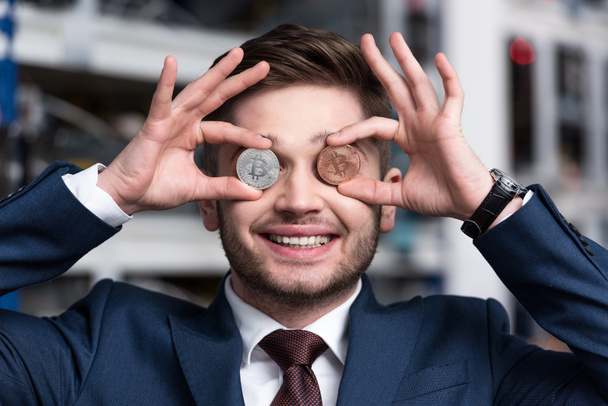 smiling young businessman covering eyes with bitcoins at cryptocurrency mining farm - Foto, afbeelding