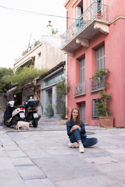 Smiling Caucasian girl. A girl with long hair in a denim suit and white sneakers sits on the floor in the street on the background colorful houses. Summertime - Valokuva, kuva