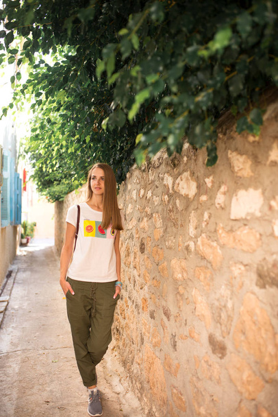 Caucasian girl in white T-shirt with fruits, green pants and gray sneakers is walking along a narrow ancient street with trees and blue shutters in the windows of the houses. - Photo, Image