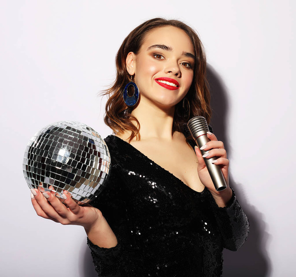 Young woman wearing black dress, holding disco ball and singing into microphone - Fotografie, Obrázek