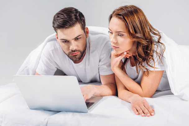 retrato de pareja enfocada usando portátil mientras descansan en la cama juntos aislados en gris
 - Foto, Imagen