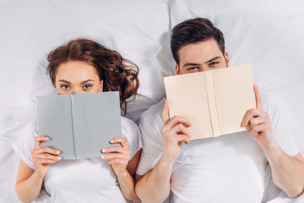 Blick aus der Vogelperspektive auf ein junges Paar, das im Bett Gesichter mit Büchern bedeckt - Foto, Bild