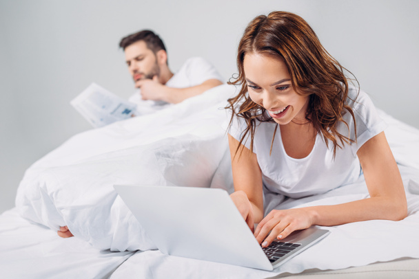 selective focus of smiling woman using laptop while lying on bed together with boyfriend isolated on grey - Φωτογραφία, εικόνα