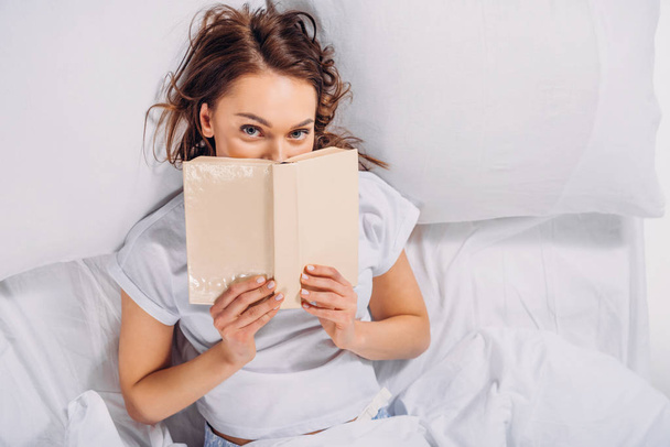 obscured view of woman covering face with book and looking at camera while lying in bed - 写真・画像