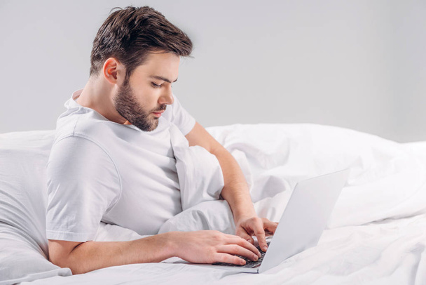 boční pohled soustředěný vousatý muž přes notebook v posteli izolované Grey - Fotografie, Obrázek