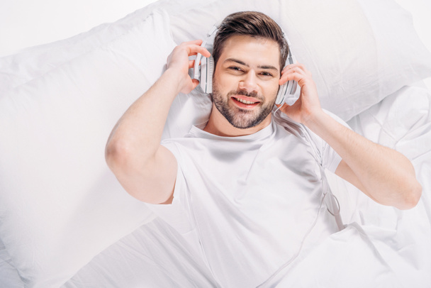 bovenaanzicht van het luisteren muziek van de mens in hoofdtelefoons glimlachen terwijl rusten op bed - Foto, afbeelding