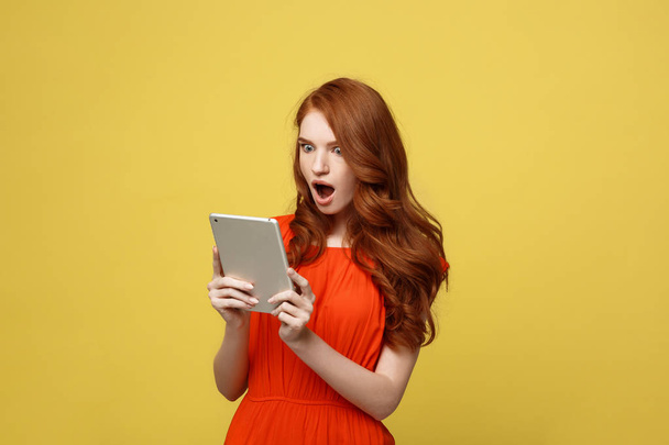 Технология и концепция образа жизни: Удивленная молодая женщина в оранжевой одежде с помощью планшетного ПК изолированы на ярком желтом фоне
 - Фото, изображение