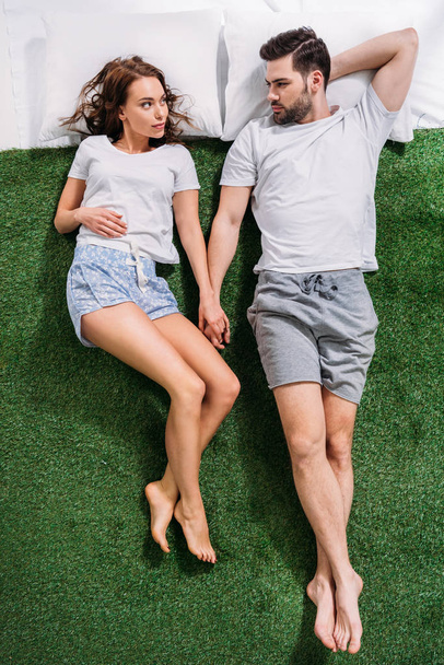 overhead view of young couple holding hands while lying on pillows on green grass - Fotografie, Obrázek
