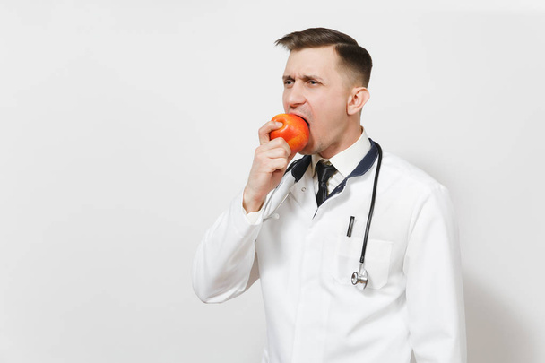 Bonito jovem médico isolado em fundo branco. Médico masculino em uniforme médico, estetoscópio mordendo comer maçã vermelha. Pessoal de saúde, conceito de medicina sanitária. Nutrição adequada. Espaço de cópia
 - Foto, Imagem