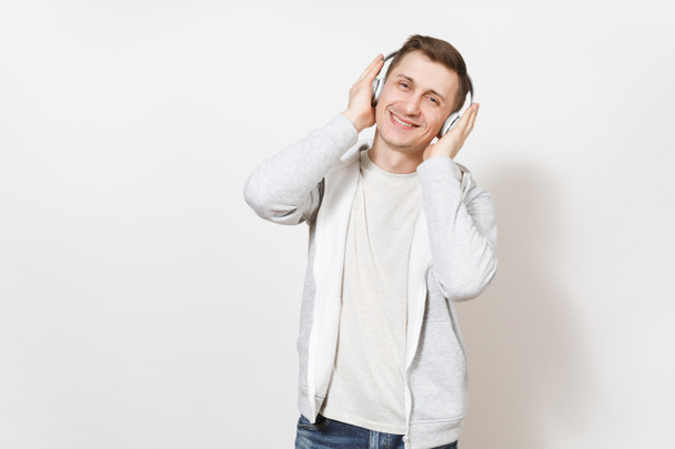 Young handsome smiling happy man student in t-shirt and light sweatshirt listening to music with white wireless headphones and puts hands to them isolated on white background. Concept of emotions - Photo, image