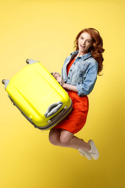 Travel and Lifestyle Concept: Portrait of a beautiful red hair fashion woman jumping and holding a bright green suitcase isolated over bright yellow background. - 写真・画像