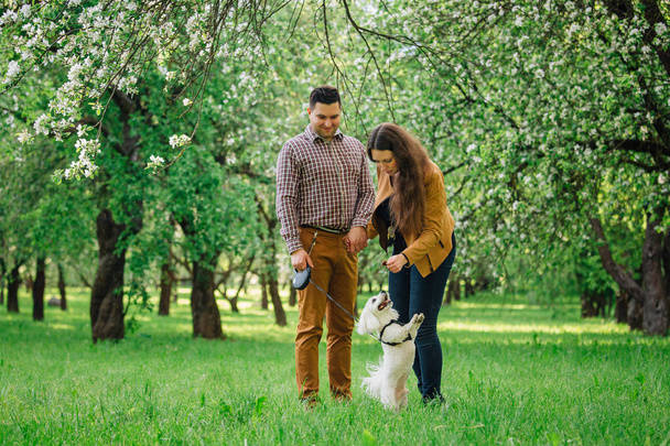 Joven pareja feliz con estilo en el amor jugando con el perrito blanco en el jardín floreciente. Chica bonita y hombre divirtiéndose con maltés en el parque
 - Foto, imagen