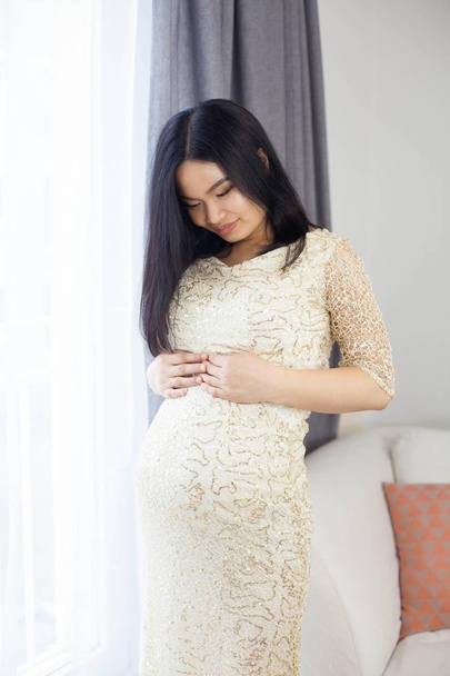Beautiful young Asian pregnant woman standing by the window and touching her tummy. Pregnancy. Health. Motherhood. A cozy portrait of a pregnant woman. Cozy. - Foto, Imagem