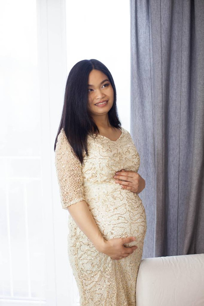 Linda jovem mulher grávida asiática em pé junto à janela e tocando sua barriga. Gravidez. Saúde. Maternidade. Um retrato acolhedor de uma mulher grávida. Aconchegante
. - Foto, Imagem
