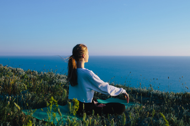 若い女性の練習ヨガで、ロータスの位置屋外。海の近くの丘でのティーンエイ ジャーの女の子リラックス。ジョギング、穏やかで瞑想のコンセプトはリラックス トレーニング、健康的なライフ スタイルと健康  - 写真・画像