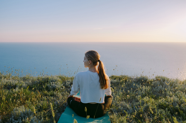 Jovem mulher praticando Yoga na posição de lótus ao ar livre. adolescente menina relaxante na colina perto do mar. Conceito de calma, meditação, corrida, treinamento, relaxamento, estilo de vida saudável e bem-estar. vista traseira
 - Foto, Imagem