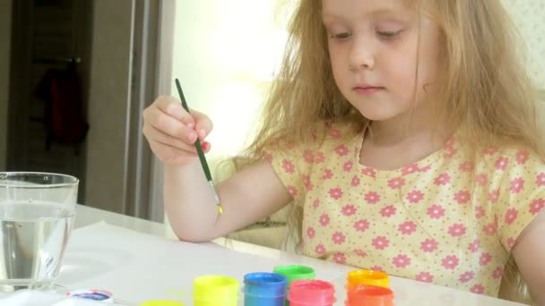 μικρή όμορφη κοκκινομάλλα κοπέλα ζωγραφίζει με paintand βούρτσα που κάθεται στο τραπέζι - Πλάνα, βίντεο