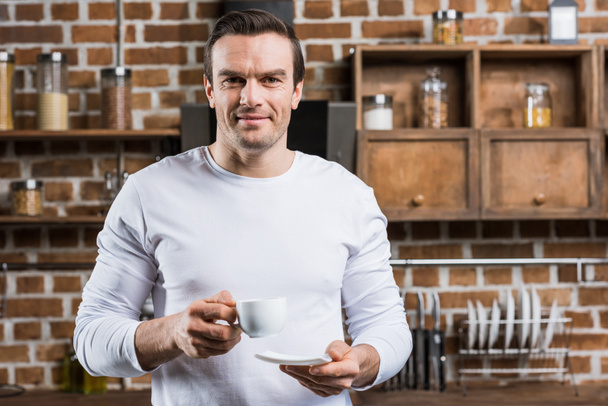 handsome man holding cup of coffee and smiling at camera in kitchen - Фото, зображення
