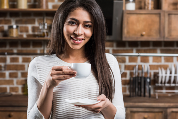 beautiful african american girl holding coffee cup and smiling at camera in kitchen - Photo, Image