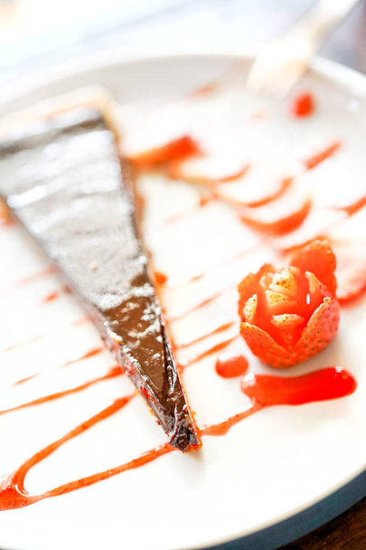 Ένα κομμάτι του κέικ σοκολάτας με φράουλες βρίσκεται σε μια λευκή πλάκα, πασπαλισμένες με μαρμελάδα. Ένα μικρό πιρουνάκι βρίσκεται πλάι-πλάι σε ένα τραπέζι με φόντο το ξύλινο. Κινηματογράφηση σε πρώτο πλάνο προβολής με αντίγραφο χώρου - Φωτογραφία, εικόνα