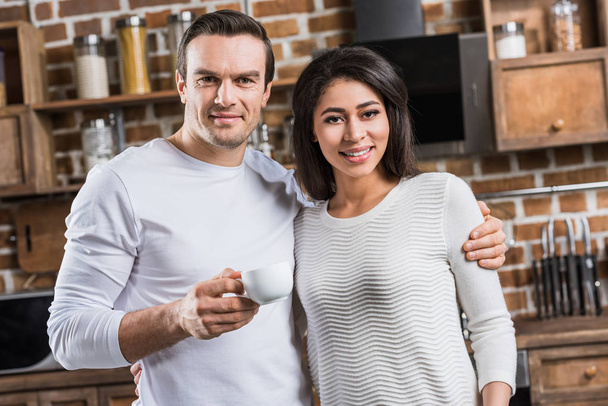 multiethnic couple embracing and smiling at camera while man holding cup of coffee in kitchen - Fotografie, Obrázek