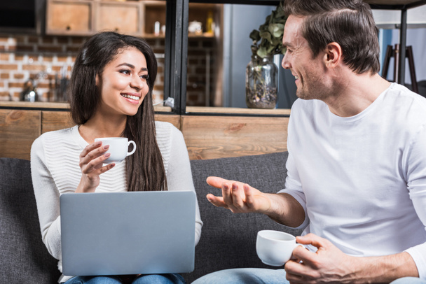 multiethnic couple smiling each other and talking while drinking coffee and using laptop at home  - Foto, Bild