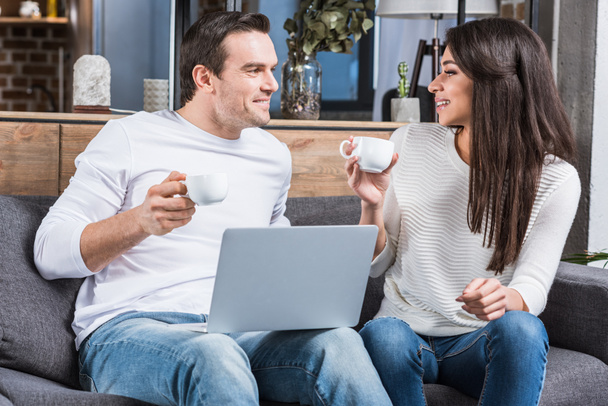 multiethnic couple holding cups of coffee and smiling each other while using laptop at home  - Fotografie, Obrázek