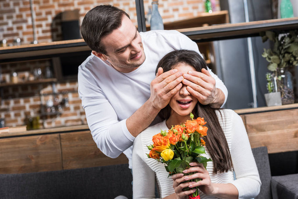 happy man closing eyes to girlfriend holding bouquet of flowers at home - Zdjęcie, obraz