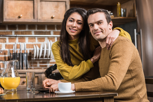 feliz joven pareja multiétnica sonriendo a la cámara mientras desayuna en casa
 - Foto, Imagen