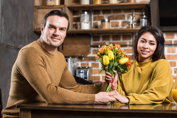 happy young multiethnic couple with flower bouquet holding hands and smiling at camera at home - Fotografie, Obrázek