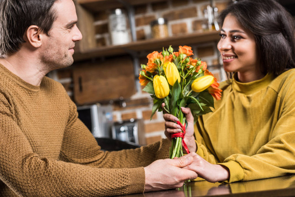 smiling young man presenting beautiful flower bouquet to happy girlfriend at home - Fotografie, Obrázek