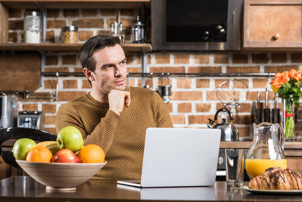 pensive man looking away while using laptop at kitchen table - Foto, Imagem