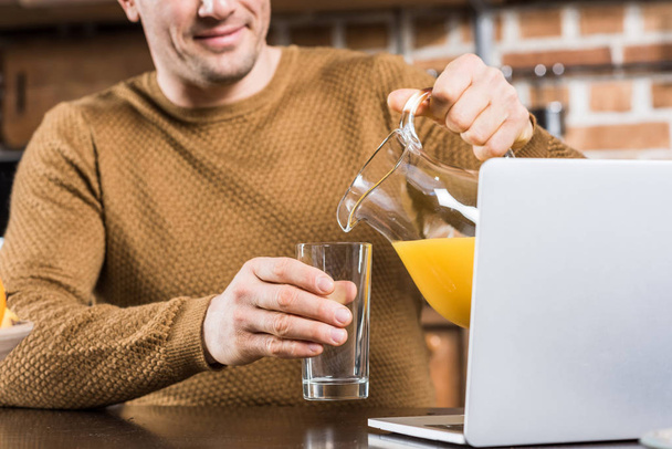cropped shot of smiling man pouring orange juice into glass while using laptop at home - Photo, Image