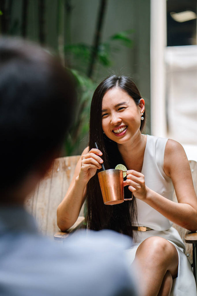 young Chinese Asian woman out on a romantic date with a man. She is drinking an alcoholic beverage while she smiles, laughs and talks with her date in a beautiful bar or cafe. - Foto, imagen
