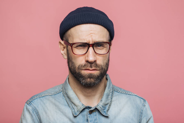 Unhappy discontent unshaven male looks with grumpy expression into camera, wears glasses black hat and stylish denim shirt, isolated over pink background. Displeased middle aged man poses indoor - Photo, Image