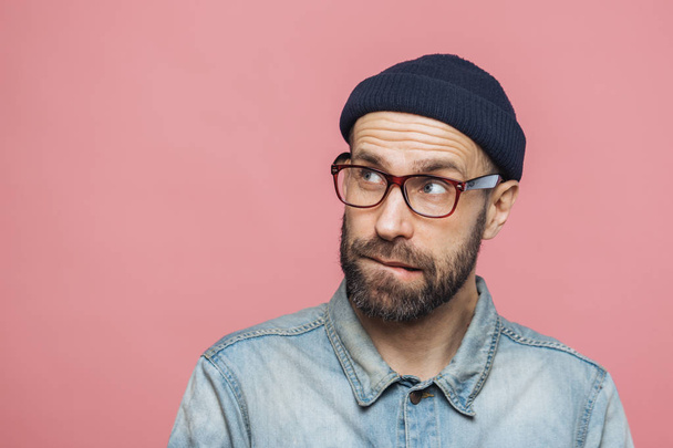 Portrait of unshaven thoughtful middle aged male with pensive expression looks upwards, wears stylish hat, eyewear and denim jacket, isolated over pink background with copy space for your text - Photo, Image