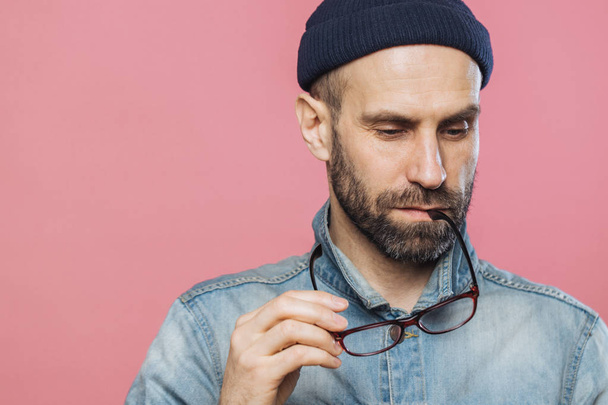 Headshot of pensive bearded man looks thoughtfully down, holds glasses, wears denim jacket and hat, isolated over pink background with copy space for your advertising content. People and thoughts - Fotó, kép