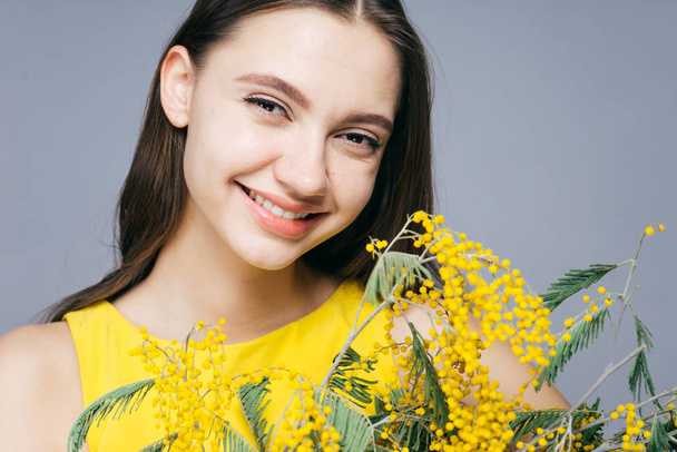 happy young girl in yellow dress holds bouquet of fragrant yellow mimosa and smiles - Foto, afbeelding