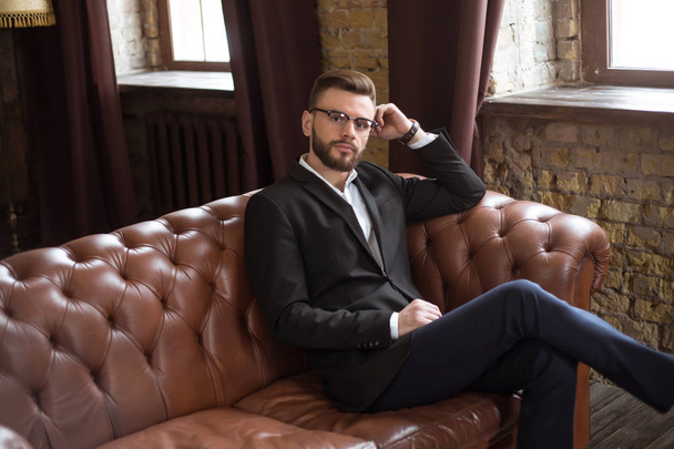 Bonito homem de negócios barbudo confiante e elegante em um terno inteligente e óculos sentados em um sofá no escritório
. - Foto, Imagem