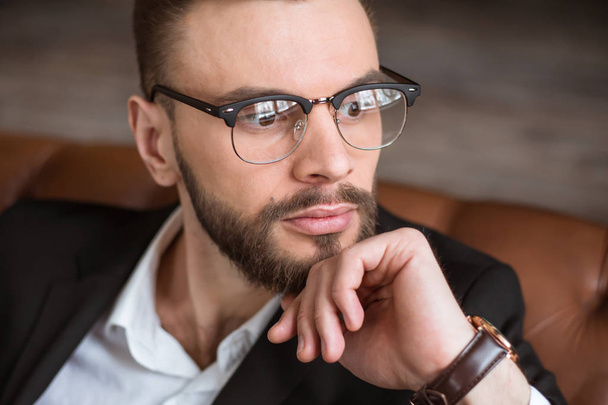 Handsome stylish confident bearded businessman in a smart suit and glasses sitting on a sofa in the office. - Фото, зображення