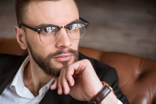 Guapo y elegante empresario barbudo con confianza en un traje inteligente y gafas sentadas en un sofá en la oficina
. - Foto, Imagen