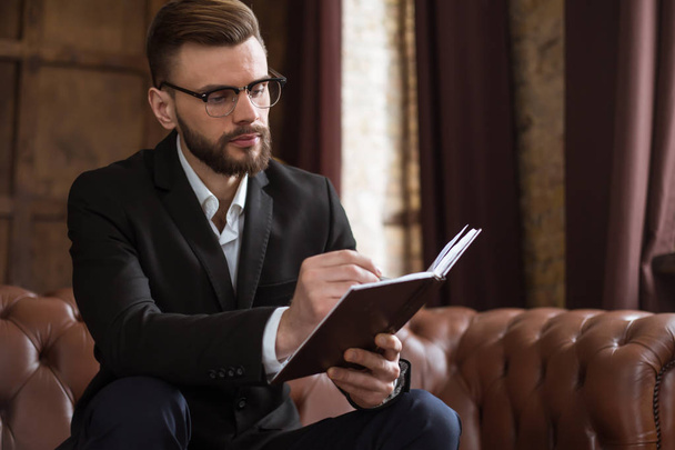 Уверенный красивый бородатый бизнесмен в умном костюме и очках читает книгу или проверяет отчеты о работе сидя на диване в офисе
. - Фото, изображение