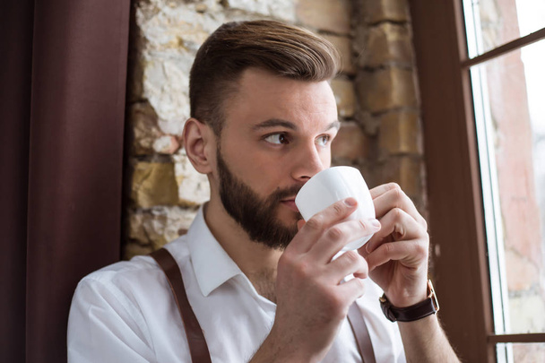 Ontspan na hard werken. Knappe bebaarde man in wit overhemd drinkt een kopje warme koffie of thee in de buurt van groot venster en weg op zoek. - Foto, afbeelding