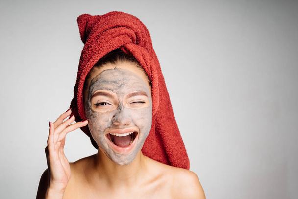 happy young girl with a red towel on her head applied a useful clay mask on her face, laughs - 写真・画像