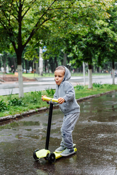Skąpane w deszczu, chłopiec w sporcie garnitur Łyżwy na skuter. Wiosenny spacer w parku miejskim, deszczowa pogoda. - Zdjęcie, obraz