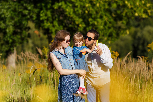 Young mother and father blowing soap bubbles and their little daughter trying to catch them. Funny kid playing outdoors. Concept on family holidays and walking at nature, parenting - Photo, Image