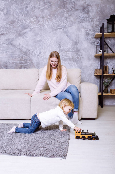 Little girl is playing with wooden toy locomotive, train. Her mother is looking on her. Blonde cute baby girl sitting at home on the carpet near the sofa. - Фото, зображення