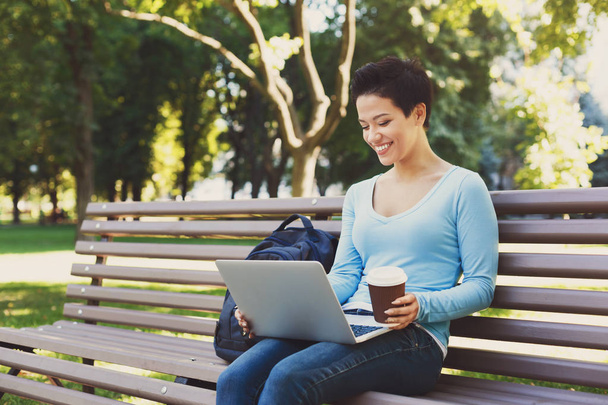 Atractiva freelancer sonriente que trabaja en una laptop y bebe café afuera
 - Foto, Imagen
